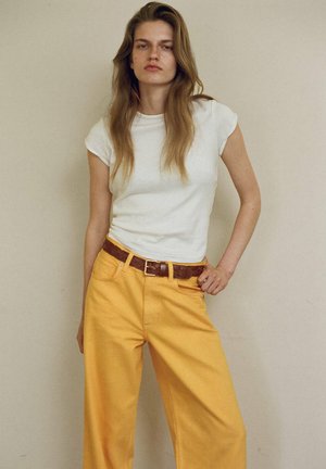 Jeune femme aux longs cheveux portant un t-shirt blanc, un pantalon large jaune et une ceinture tressée marron, debout avec une main sur la hanche contre un mur uni.