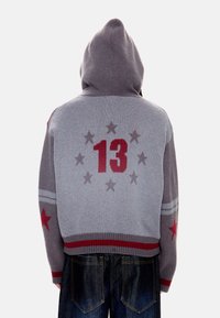 Άτομο που φοράει γκρι φούτερ με κουκούλα με κόκκινα αστέρια στα μανίκια και τον αριθμό 13 περιβαλλόμενο από αστέρια στην πλάτη, συνδυασμένο με σκούρο τζιν.