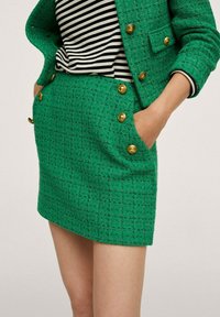 Blazer en tweed texturé vert avec boutons dorés, porté sur un haut rayé noir et blanc et une jupe texturée verte assortie avec poches.