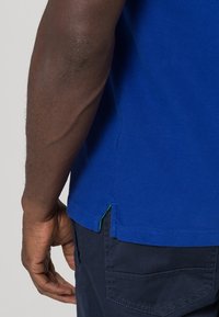 Kortärmad blå bomullströja med en sidöppning och grön accent. Strukturerad tyg med minimal sömdetalj vid fållen.