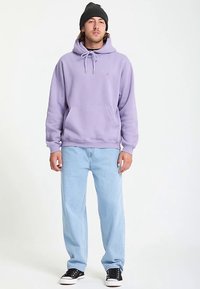 Paarse hoodie met voorzak en capuchon met trekkoord, gecombineerd met lichtblauwe spijkerbroek en zwarte sneakers. Het materiaal lijkt zacht en comfortabel.