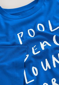 Modrá látka s bielym textom, kde sú viditeľné časti slov "POOL," "BEAC," a "LOUN", naznačujúce neformálny dizajn trička.