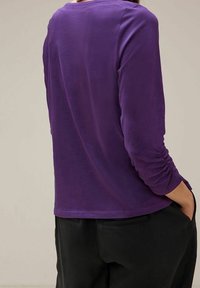 Lila långärmad topp med rund halsringning och veckade detaljer på ärmarna. Tyget verkar slätt och något lätt.