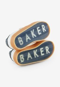Valkoiset lenkkarit, joissa on siniset kumipohjat, herringbone-kuviointi ja valkoinen "BAKER"-teksti. Pohjien ruskea kumireuna.