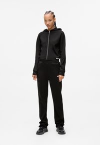 Giovane con capelli intrecciati, indossa una felpa nera con zip, pantaloni della tuta neri abbinati e sneakers nere, in piedi su uno sfondo bianco semplice.