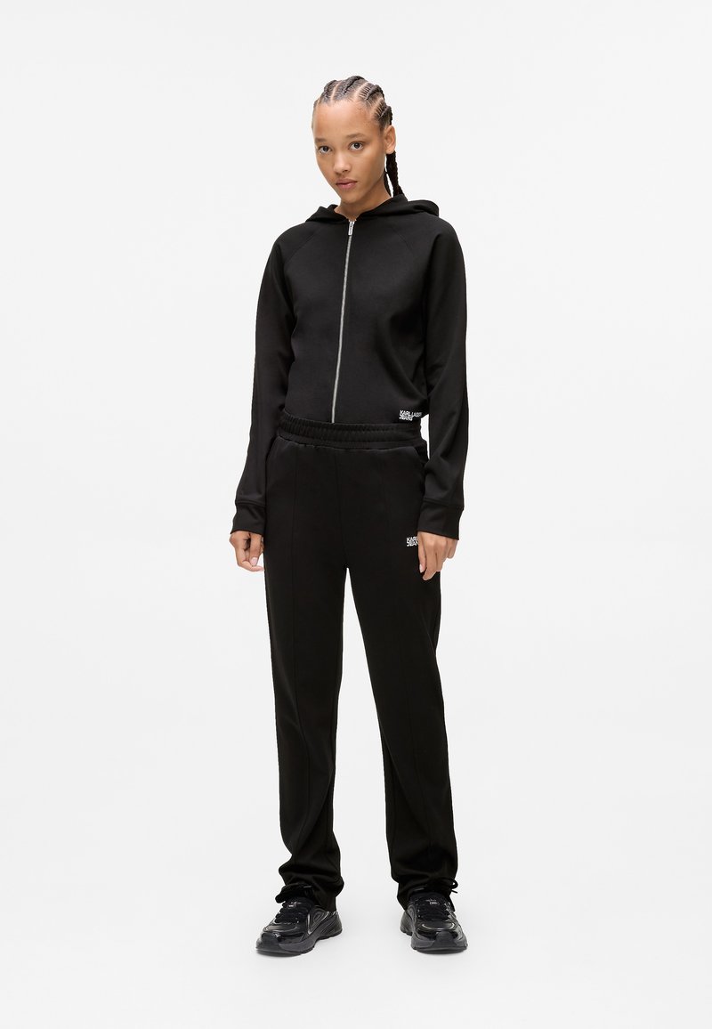 Giovane con capelli intrecciati, indossa una felpa nera con zip, pantaloni della tuta neri abbinati e sneakers nere, in piedi su uno sfondo bianco semplice.
