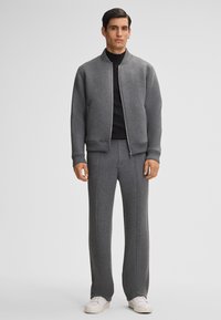 Mann steht mit einer grauen Zip-Jacke, passenden grauen Hosen, schwarzem Shirt und weißen Sneakers vor einem einfachen hellgrauen Hintergrund.