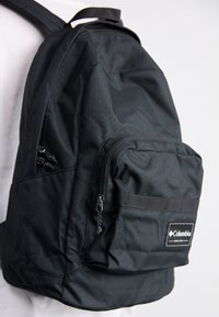 Czarna plecak wykonany z trwałego materiału, z regulowanymi paskami, przednią kieszenią na zamek oraz naszywką z logo marki. Posiada gładką strukturę.