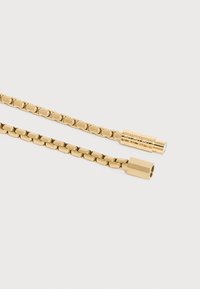 Δύο συνδεδεμένα άκρα χρυσής αλυσίδας για κολιέ, το ένα με κυλινδρικό κούμπωμα με εγχάρακτο σχέδιο και το άλλο με τετράγωνο κούμπωμα.