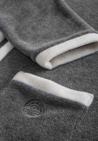 Gray fleece garment with white inner lining, featuring a contrast hem and a circular logo detail. Soft texture with pronounced seams.