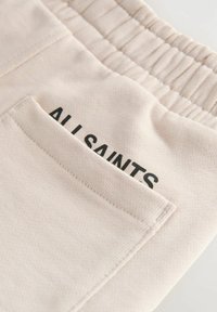 Helle beige Baumwollhose mit elastischem Bund und einer Gesäßtasche mit schwarzem "ALL SAINTS"-Druck. Glatte Textur, lässiges Design.