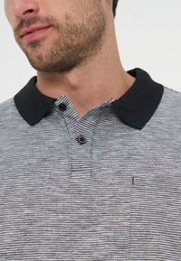 Άνδρας που φοράει γκρι και άσπρη ριγέ πόλο μπλούζα με μαύρο γιακά και δύο κουμπιά, μερική άποψη του κάτω μέρους του προσώπου και του λαιμού του.