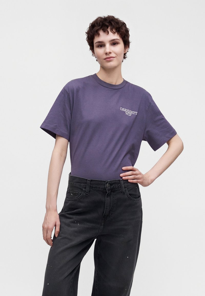 Giovane con capelli corti e ricci che indossa una t-shirt viola Carhartt WIP e jeans neri, in piedi con una mano sui fianchi contro uno sfondo semplice.