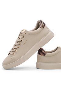 Baskets basses beiges avec lacets beiges, œillets dorés et détail du talon à motif tigre, avec un logo de la marque sur le côté.