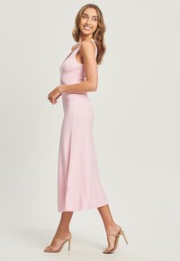 Abito midi senza maniche di colore rosa con corpetto aderente e gonna svasata. Realizzato in tessuto morbido, presenta slittini laterali. Indossato con tacchi nudi con bande.