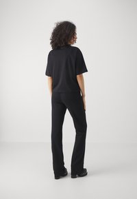 Kvinna med lockigt hår som bär en svart t-shirt med kort ärm, svarta byxor och plattformsskor, står mot en vit bakgrund.