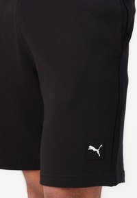 Shorts negri din bumbac, cu o textură netedă, croi până la genunchi și un logo alb Puma pe o etichetă neagră. Design simplu, fără modele.