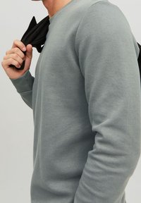 Sudadera gris de cuello redondo hecha de un tejido suave, con mangas largas y puños acanalados. Un artículo negro está colgado sobre el hombro del modelo.