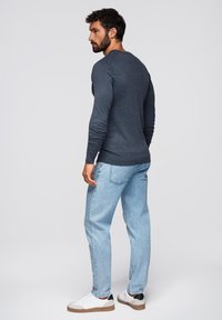 Maglietta grigia scura a maniche lunghe, colletto tondo; abbinata a jeans blu chiaro; sneakers bianche con dettagli marroni. Modello di profilo, mostrando la vista posteriore.