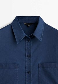 Chemise bleu marine à boutons avec col pointu et deux poches poitrine avant, présentée à plat sur un fond blanc.