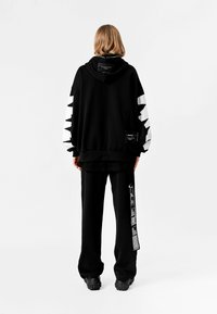 Sudadera oversized negra con detalles gráficos en blanco en las mangas, combinada con pantalones negros que tienen una franja estampada. Textura suave, diseño casual.