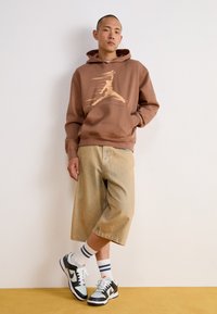 Sweat à capuche marron avec un imprimé graphique, short en denim beige, baskets noires et blanches, et chaussettes rayées, le tout sur un fond neutre.