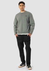 Ung mand står med hænderne i lommerne iført en grå sweater, sorte bukser og sort-hvide sneakers foran en ensfarvet grå baggrund.
