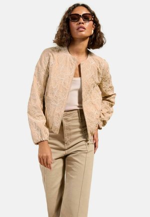 Vrouw die een beige bomberjack met bloemenborduursel draagt, een licht tanktopje, hoge beige broek, zonnebril en gouden creolen.