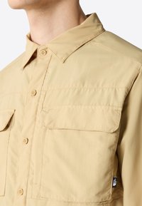 Camicia button-up color khaki con colletto, due tasche sul petto e una trama sottile. Presenta un'etichetta con logo sul lato per il branding.