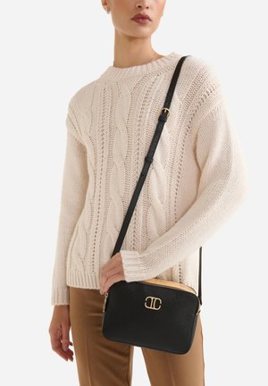 Donna che indossa un maglione crema a trecce e pantaloni marroni, con una borsa a tracolla nera con logo dorato e tracolla regolabile.