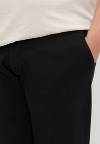 Black pants made of cotton, featuring a smooth texture and side pockets, shown with a light-colored shirt above the waist.