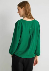Una donna con capelli castani lisci indossa una blusa verde a maniche lunghe con texture e pantaloni neri, di profilo con le mani in tasca.