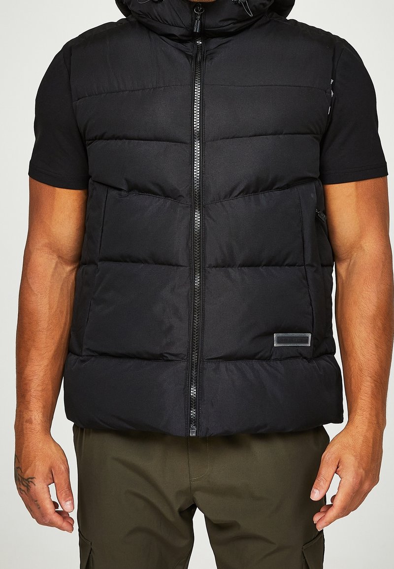 Černá prošívaná vesta s kapucí, s předním zipem, dvěma postranními kapsami a texturovaným povrchem. Obsahuje malou značkovou nášivku.