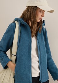Veste en polaire zippée en teal avec une finition texturée, dotée d'une capuche, de poches latérales et d'une sangle de couleur claire provenant d'un sac à main.