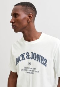 Hvid T-shirt med "JACK & JONES" i blå tekstureret skrifttype. Rund hals, korte ærmer og afslappet pasform med trykte detaljer.
