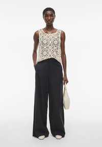 Top de tirantes de encaje en beige con recortes florales, combinado con pantalones anchos negros y zapatos de punta beige. Sosteniendo un pequeño bolso blanco.