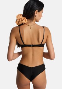 Μαύρο μπικίνι με λεπτές τιράντες και κλείσιμο στο πίσω μέρος. Πορτοκαλί υφασμάτινο scrunchie με υφή γκίνγχαμ στα μαλλιά. Ορατό χρυσό κολιέ αλυσίδα.