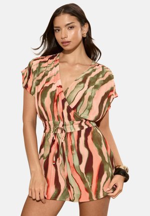 Žena nosí krátky overal s krátkym rukávom a výstrihom do V s ružovými, zelenými a hnedými vlnitými pásmi, v páse zúžený sťahovacím pásikom.