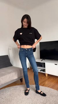 Czarny t-shirt z krótkim rękawem z różowym sercem, do tego niebieskie obcisłe dżinsy i czarne baleriny. Jasne tło z dywanem i meblami.