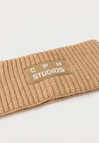 Bentiță tricotată în maro deschis, cu textură nervurată. Prezintă o etichetă dreptunghiulară bej cu text alb inscripționat "CPH Studios".