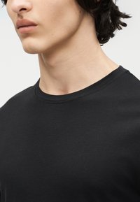 Junge Person mit kurzen dunklen Haaren, die ein schlichtes schwarzes T-Shirt mit Rundhalsausschnitt trägt und mit dem Gesicht nach links vor hellem Hintergrund steht.