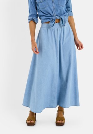 Femme portant une jupe longue en denim bleu clair, une chemise nouée en denim bleu avec manches retroussées, et des sandales compensées marron.