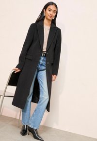 Lange zwarte jas met uitgesneden revers, twee voorzakken, knoopsluiting, gecombineerd met lichtblauwe denim jeans en zwarte puntige hakken.