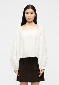 Jeune femme aux cheveux noirs et raides portant un chemisier blanc à manches longues avec des détails en dentelle et une jupe marron foncé avec des poches à l'avant.