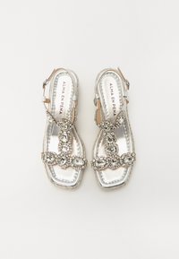 Sandali slingback argento con suola piatta, adornati con grandi strass in un motivo intricato, caratterizzati da una finitura metallica liscia.