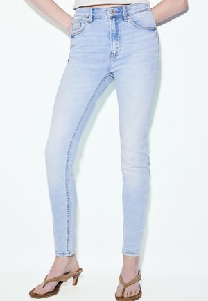 Hellblaue High-Waist Skinny-Jeans, getragen mit braunen Sandalen mit Absatz, zeigen die Beine von der Taille bis zu den Füßen vor weißem Hintergrund.