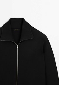 Veste noire tricotée zippée avec un col haut côtelé et une fermeture éclair argentée sur fond blanc.
