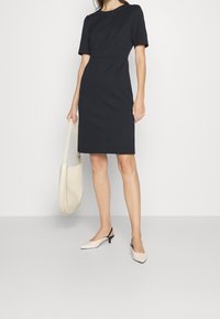 Robe noire à manches courtes avec une silhouette ajustée et une couture à la taille, associée à un sac clair et des chaussures à bride arrière bicolores.