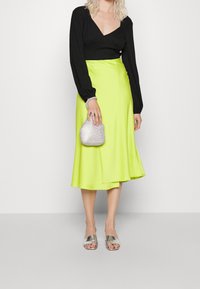 Svart långärmad topp, lysande neongrön satäng midi-kjol, silverfärgad handväska med texturerad yta och metalliska slidesandaler.