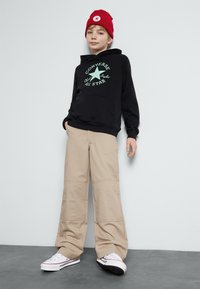 Fekete kapucnis pulóver zöld "Converse All Star" felirattal, bézs bő nadrággal és fehér tornacipővel, kiegészítve egy piros kötött sapkával.